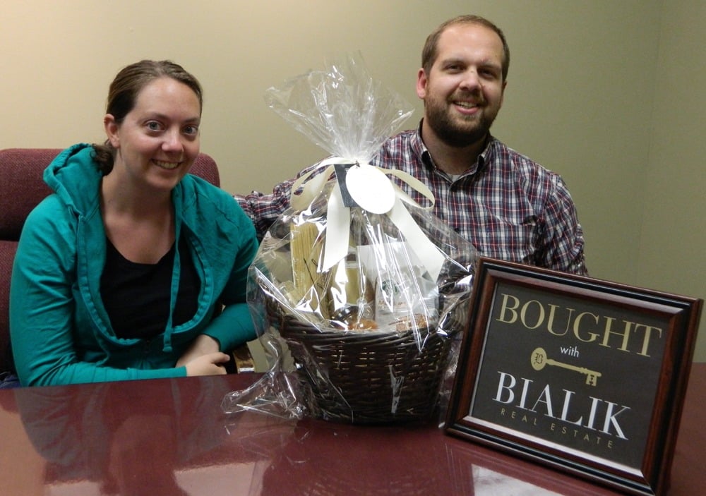 Dustin and Katie smile beside a gift basket and a Bialik Real Estate “Bought” sign.