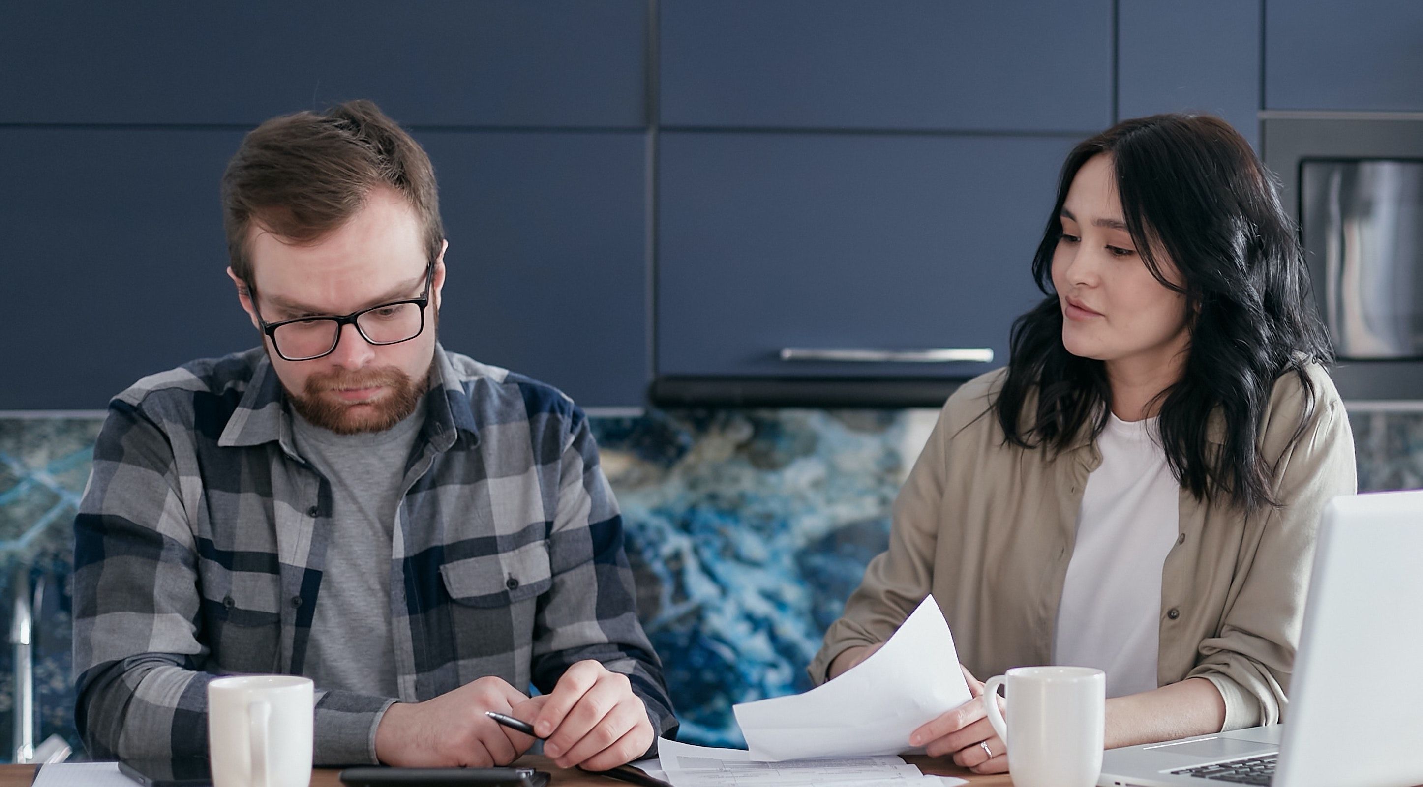 A couple reviews paperwork together at a kitchen counter, with a laptop and mugs nearby.