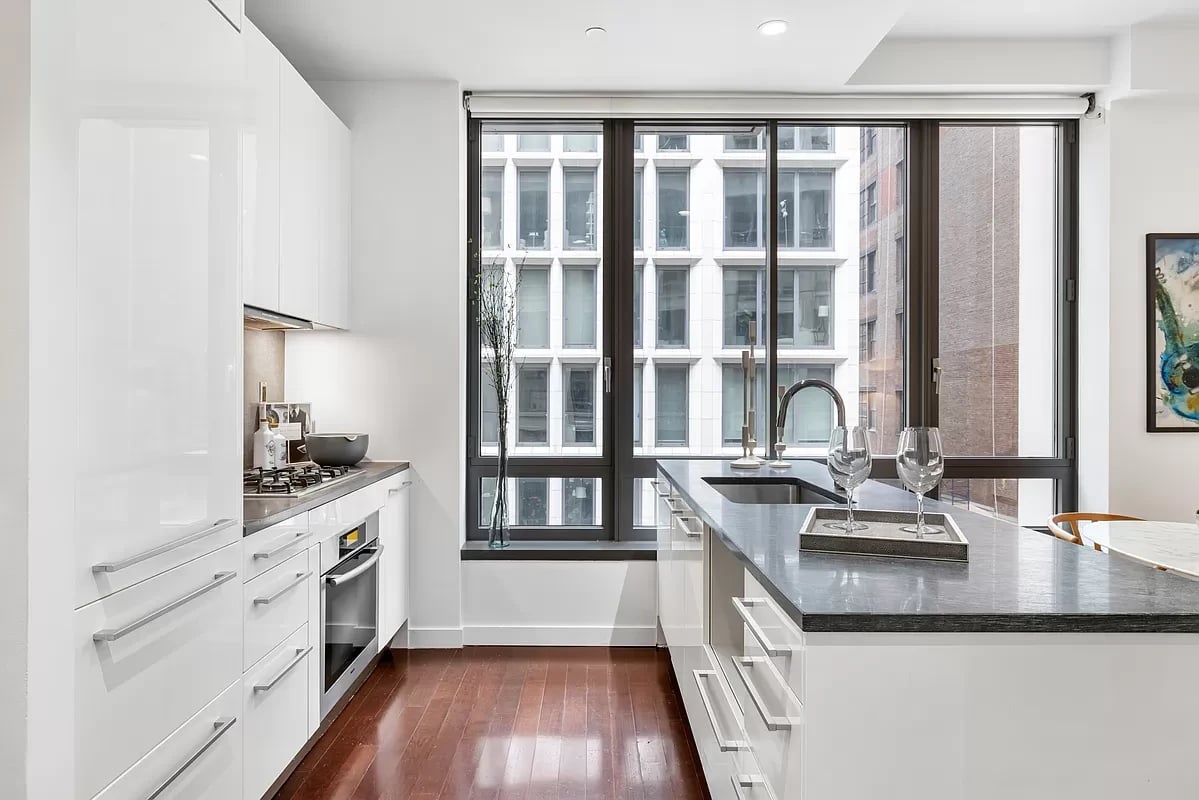 Modern white kitchen with a dark stone island, hardwood floors, and large city-facing windows.