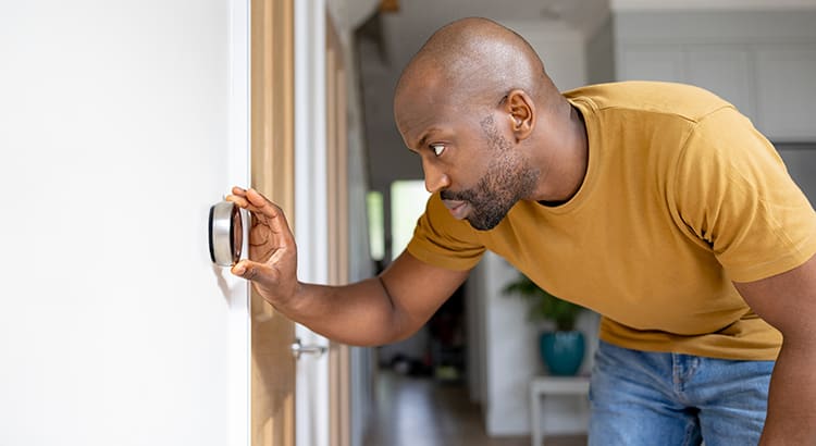 A man adjusting a thermostat on the wall. He is wearing a yellow shirt and blue jeans.