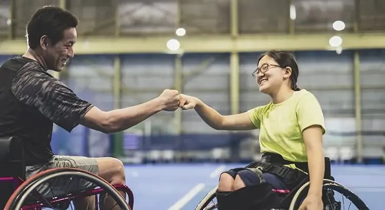 Two people in wheelchairs giving each other a high-five in a sports setting, indicating a moment of encouragement and camaraderie.