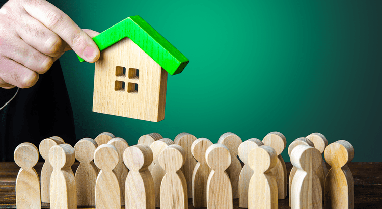 A hand placing a small house model with green roof among wooden figures, representing themes of home buying or real estate market.