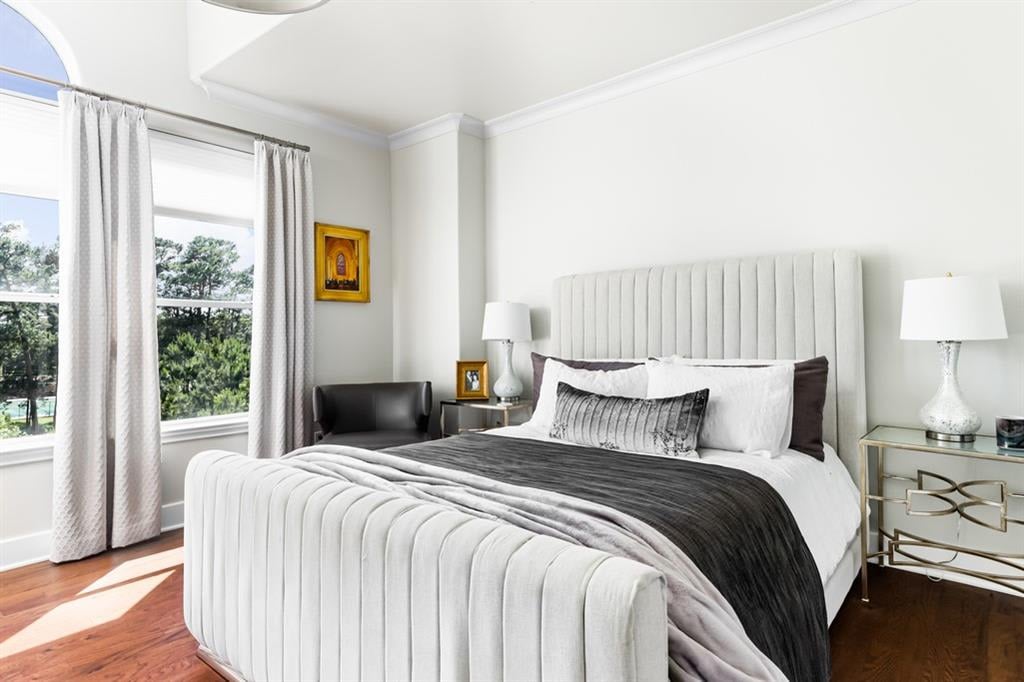 Bedroom with a tall upholstered bed, gray and white bedding, hardwood floors, and sunny windows.