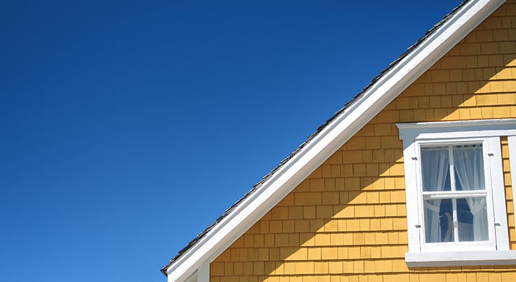 Yellow-shingled house with a white-trimmed window and steep roofline against blue sky.