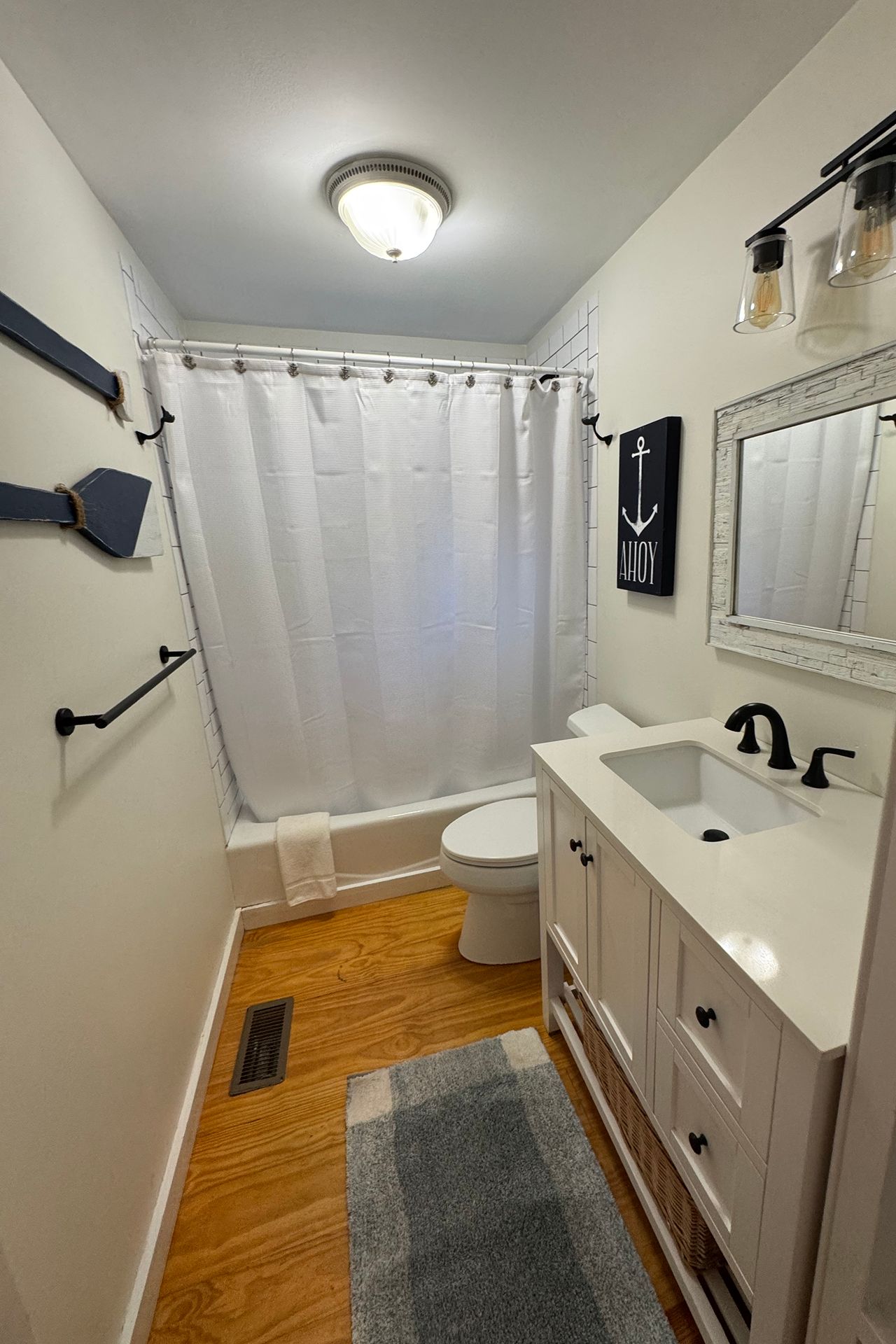 Bathroom with a white vanity and bathtub behind a shower curtain, featuring nautical wall decor and wood flooring.