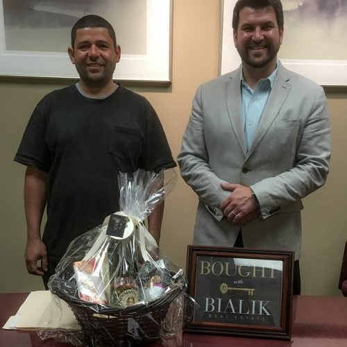Two men pose with a gift basket and a framed “Bought with Bialik Realty” sign.