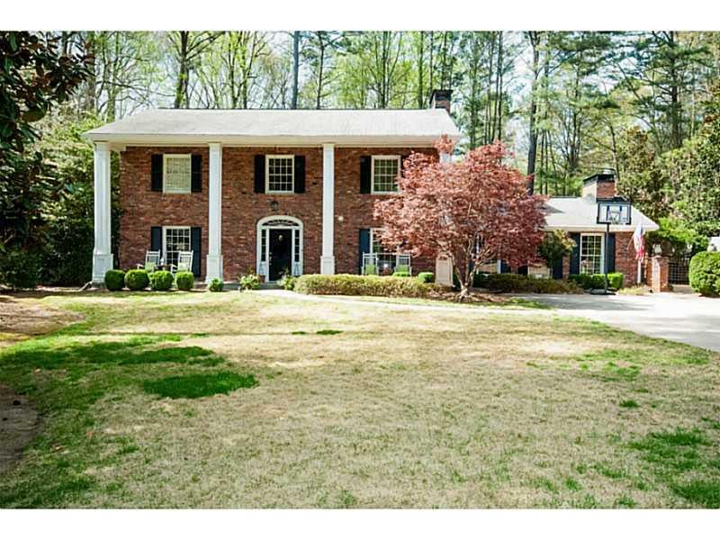 Red-brick colonial home with white columns, broad front lawn, and driveway basketball hoop.