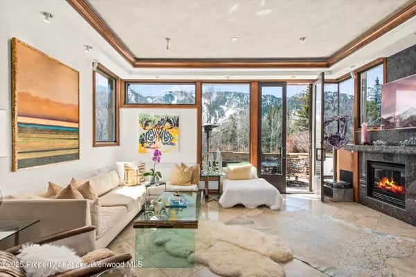 Living room with a cream sectional, lit fireplace, and floor-to-ceiling windows overlooking mountain peaks.