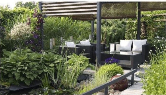 Garden patio with wicker seating beneath a pergola, surrounded by lush plants and ornamental grasses.