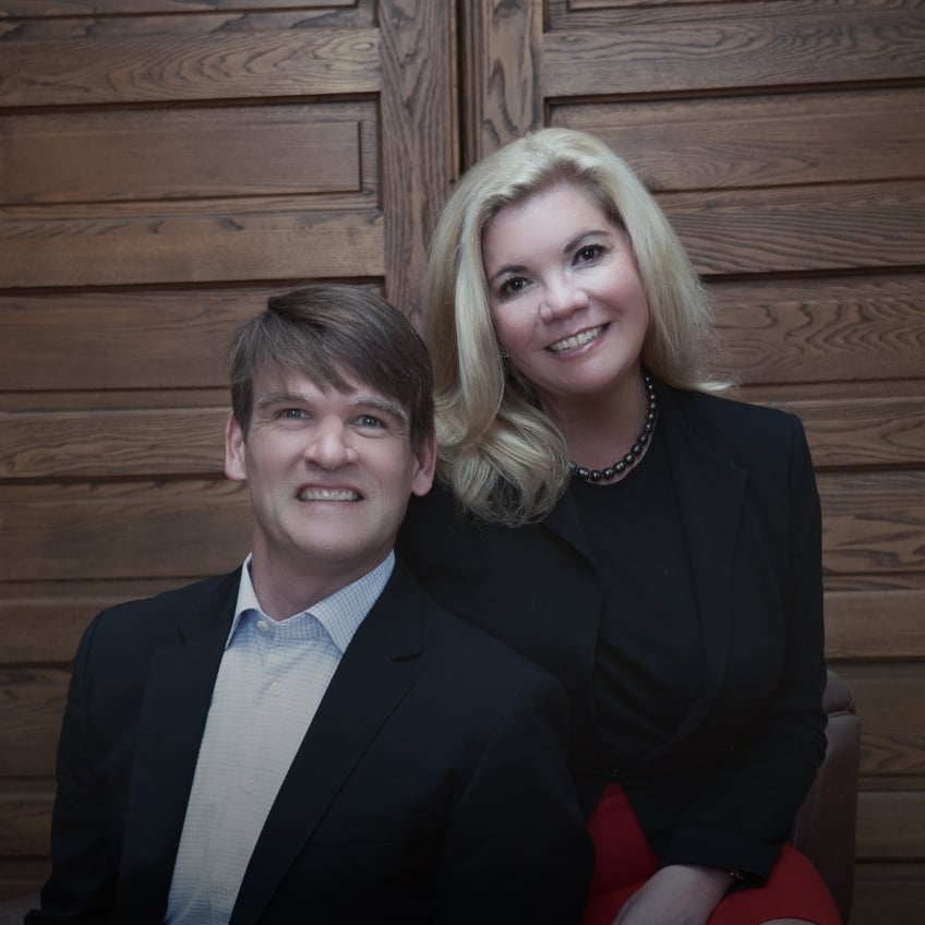 Two adults in business attire pose together against a paneled wood wall.