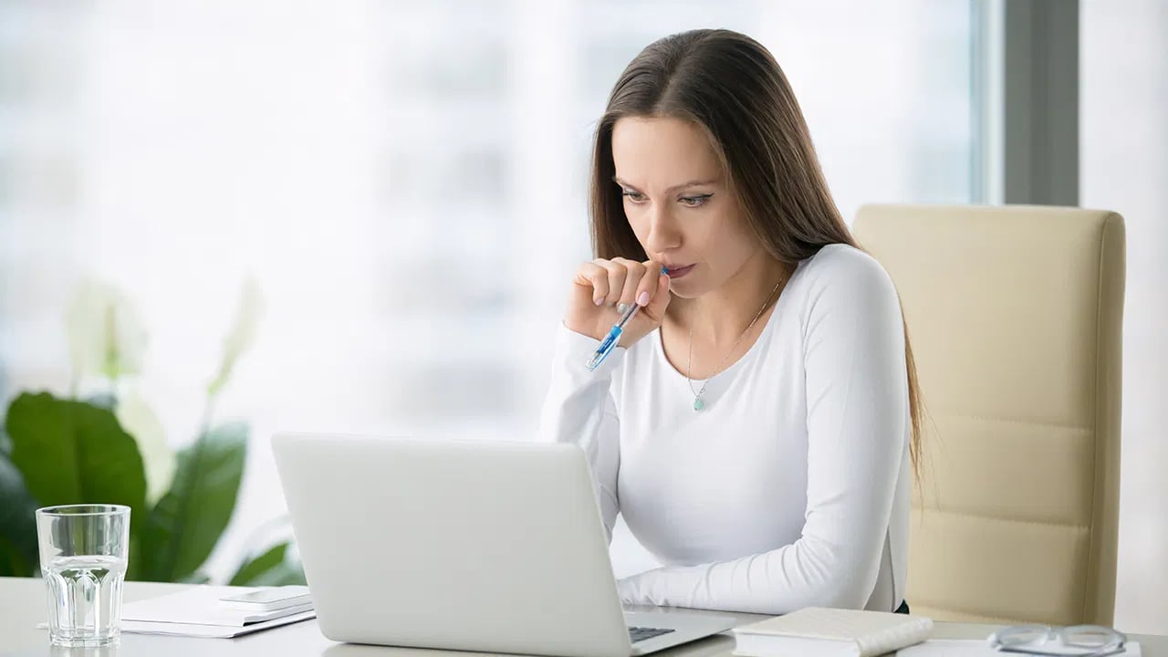 Woman thoughtfully reviewing a laptop at a desk, holding a pen with papers and a glass of water nearby.