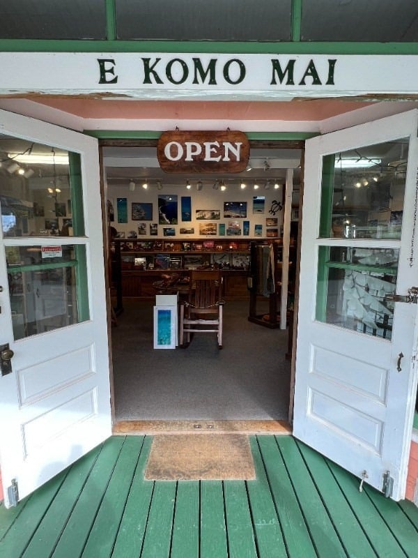 Open shop entrance with green trim, display cases, and an “E Komo Mai” sign.