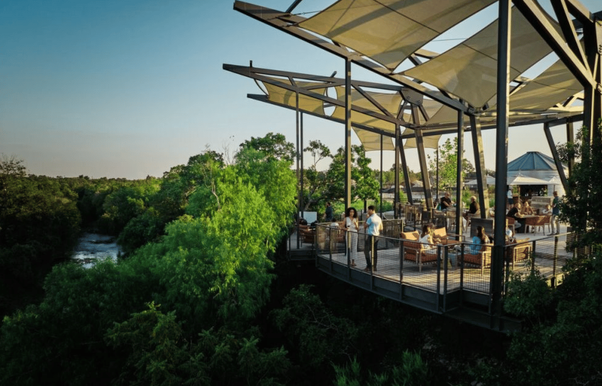 Outdoor dining terrace with shade canopies overlooking a wooded creek.