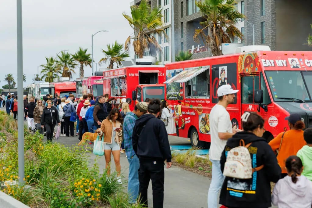 Crowd of people walking along street lined with red food trucks, palm trees and modern residential buildings visible in background