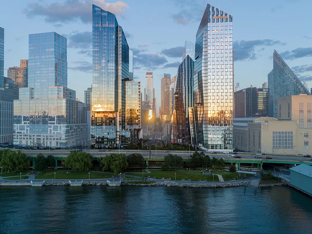Waterfront skyscrapers at sunset, with sunlight glowing between the towers above a riverside park.
