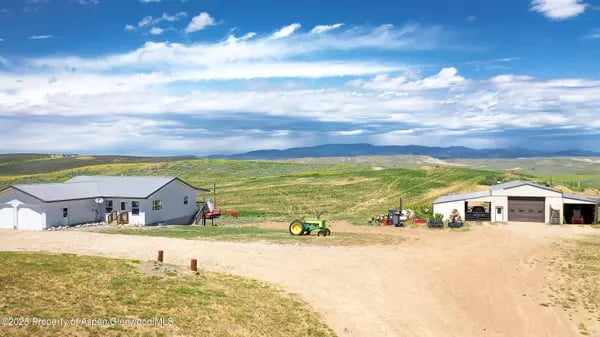 Rural property with two buildings and a tractor, surrounded by open fields and distant mountains.