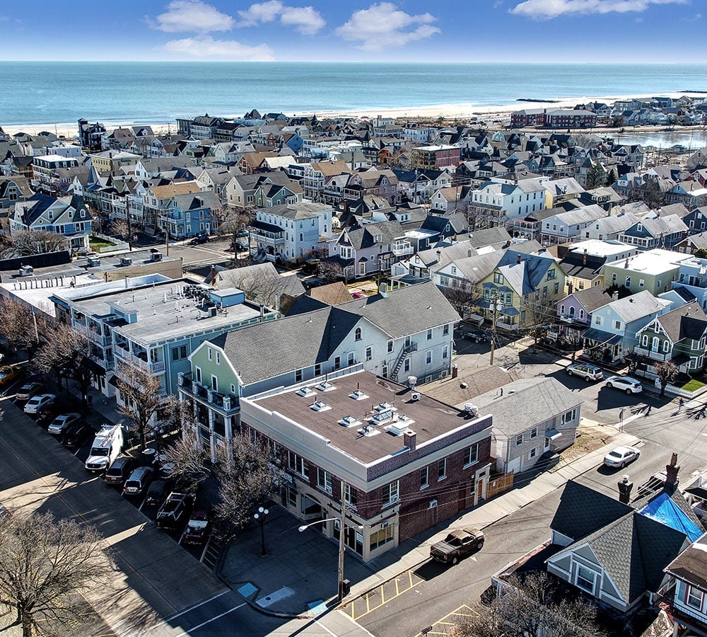 Aerial view of a coastal neighborhood and ocean, with a brick corner building in the foreground.