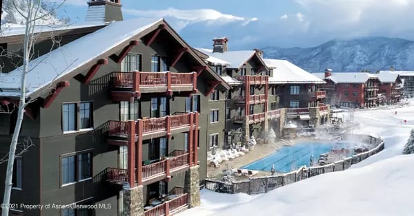 Aspen condominiums with red-brown balconies and an outdoor pool, surrounded by snow and mountain peaks.