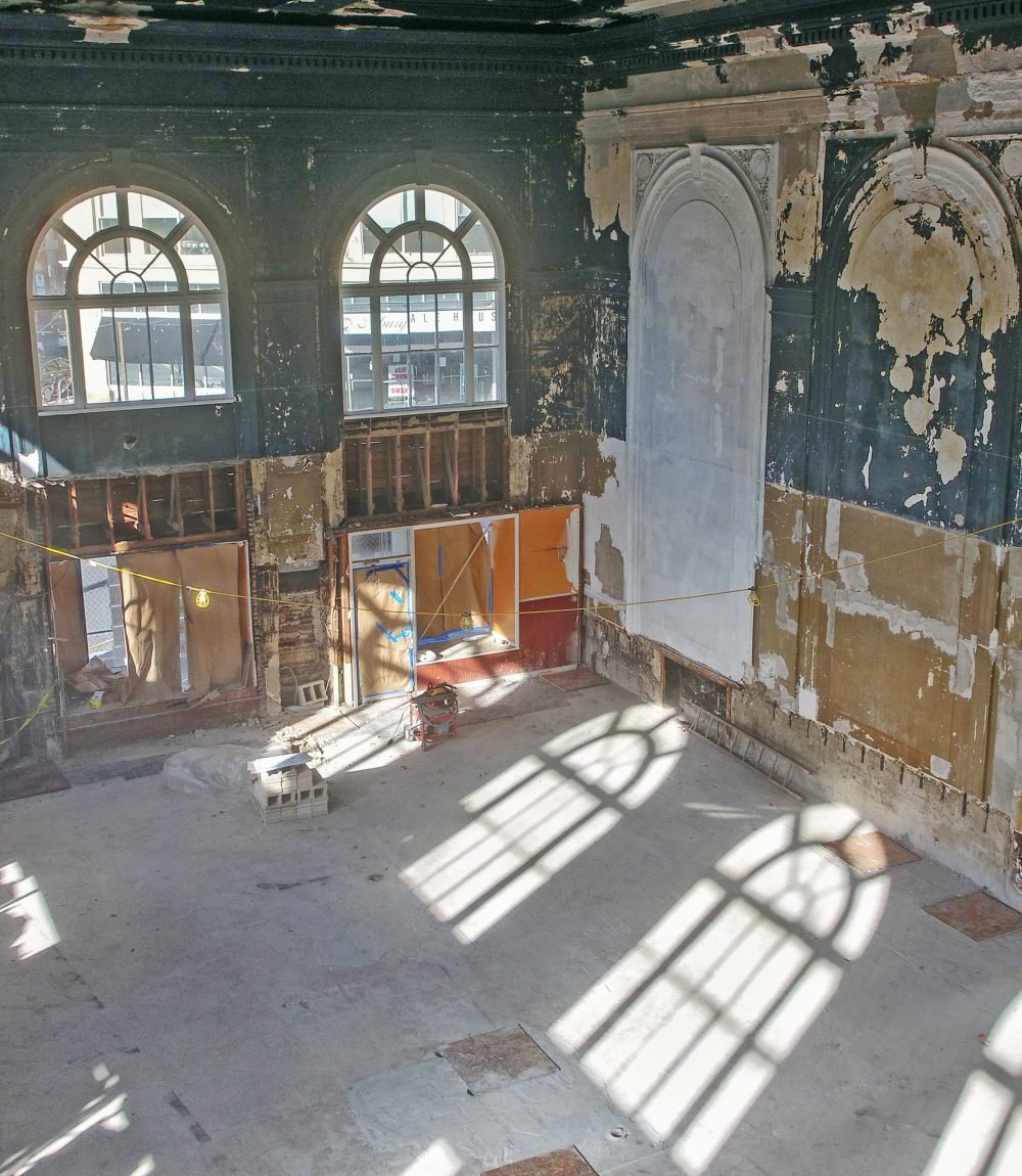 Historic interior under construction with arched windows, ornate wall niches, and sunlight across the bare floor.