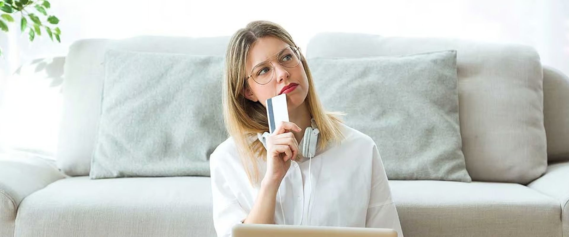 Woman thoughtfully holding a bank card while sitting in front of a light sofa.