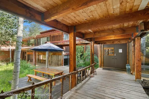 Covered timber walkway beside a modern glass-fronted building, overlooking a courtyard with a picnic table.