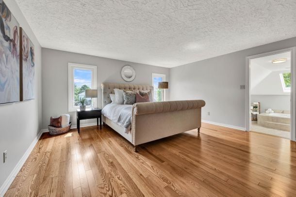 Spacious bedroom with a tufted bed, warm hardwood floors, and an adjoining bathroom doorway.