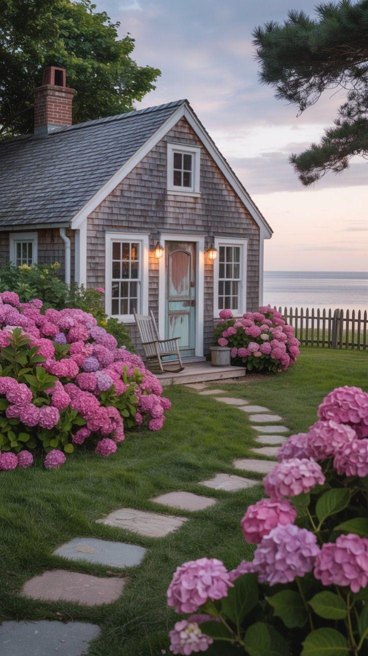 Shingled cottage beside the water, framed by pink hydrangeas and a stone garden path.