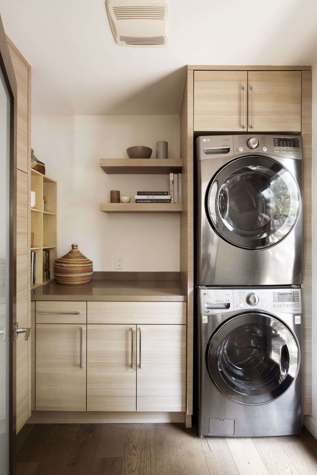 Laundry room with stacked stainless-steel washer and dryer beside light wood cabinetry and open shelves.
