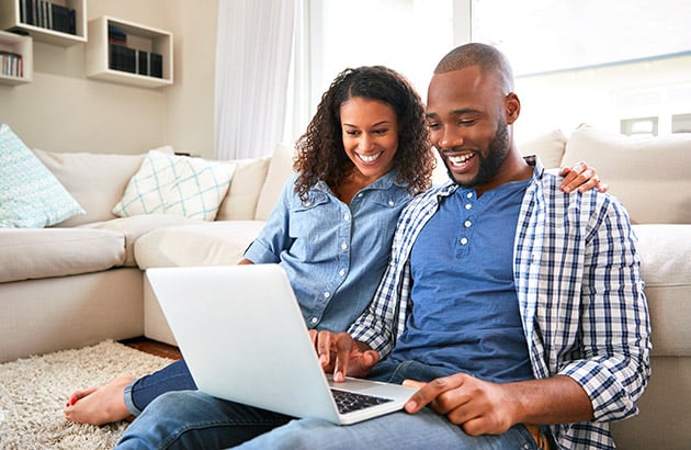A smiling couple uses a laptop together on the floor of a bright living room.