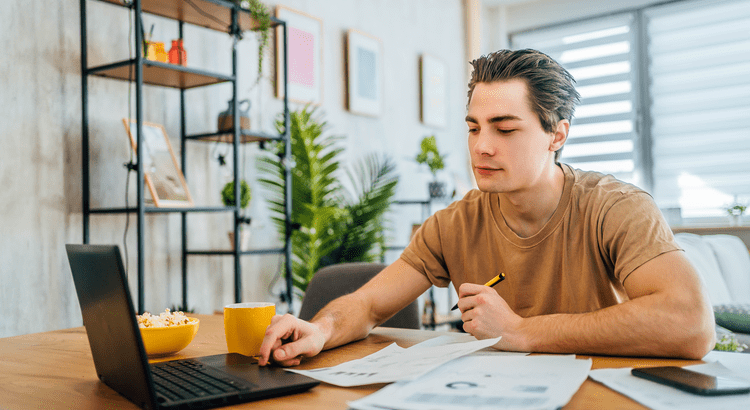 Young man working at a table with a laptop and papers, holding a yellow pencil. A mug and plant are nearby. Bright, homey atmosphere.