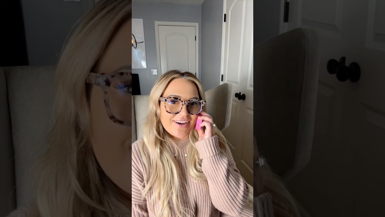 Blonde woman in patterned glasses and a blush sweater holds a pink phone while seated.