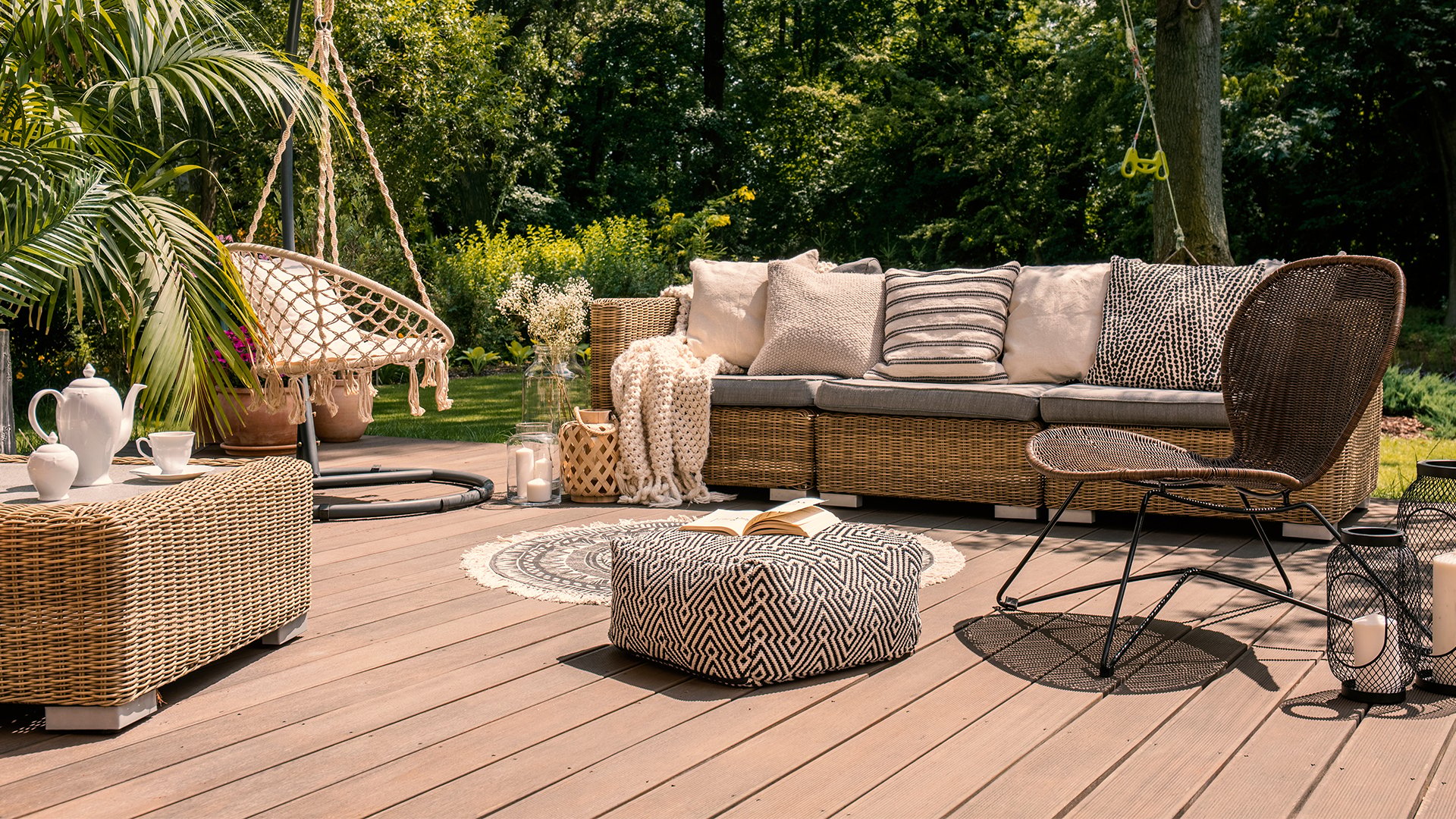 Outdoor deck with a wicker sectional, patterned cushions, and a hanging chair amid lush greenery.