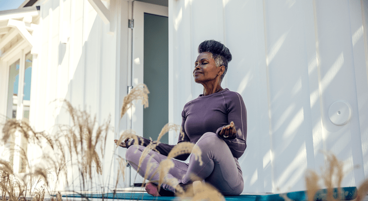 Elderly woman in purple activewear meditates outdoors, sitting cross-legged on a mat. She appears peaceful, eyes closed, with sunlight casting shadows.
