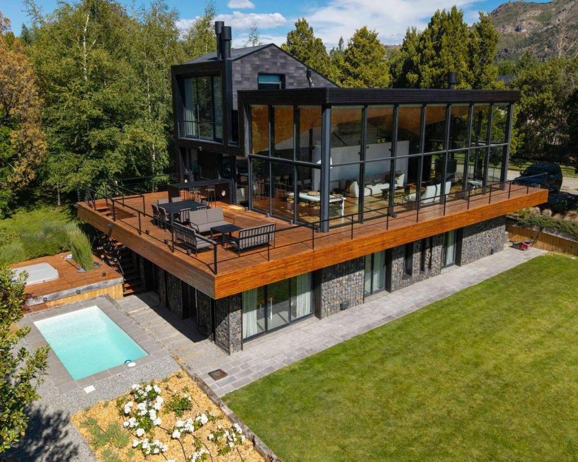 Modern glass-and-stone home with a wraparound wood deck, swimming pool, and wooded hills beyond.