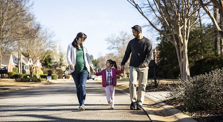 A family walking together outside. There are trees and a pathway, indicating a suburban or park setting. The image conveys a sense of family and outdoor activity.