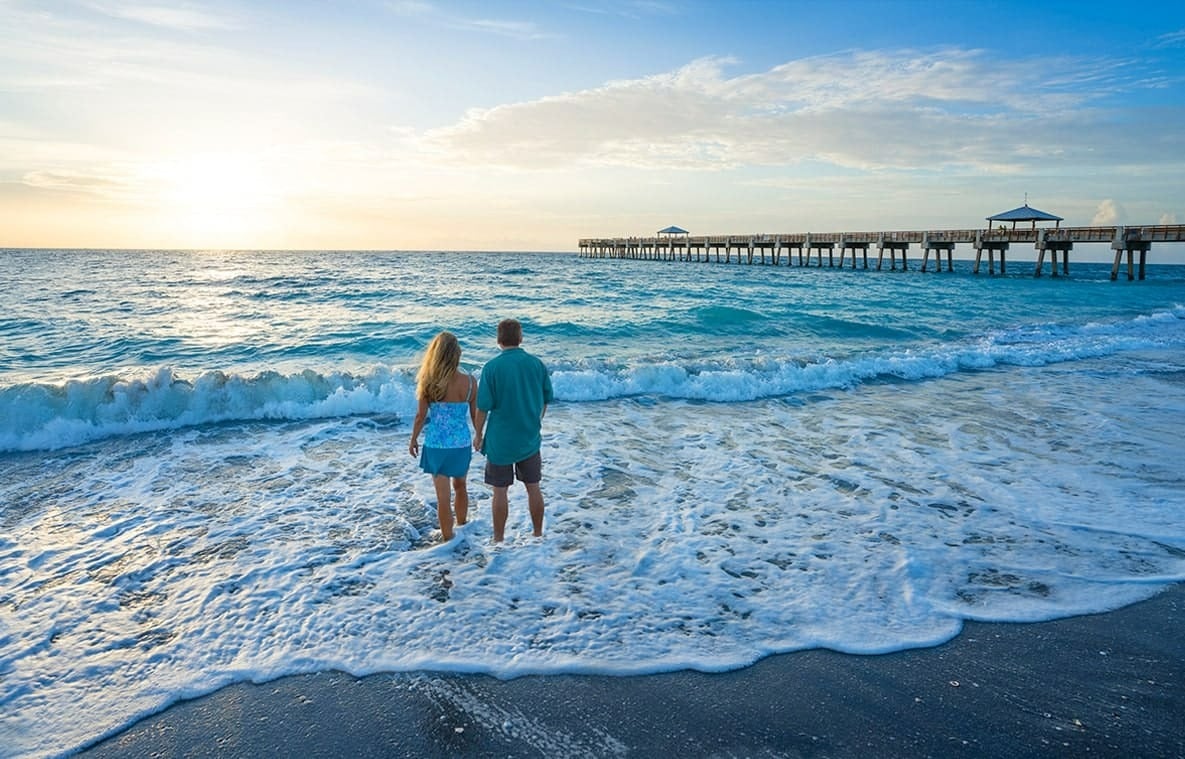 Couple watches waves at the beach beside a long pier at golden hour.