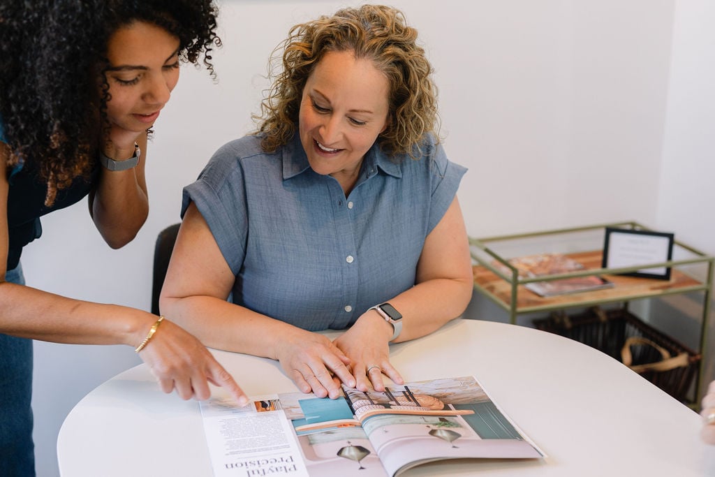 Two women review an open brochure at a table, with one pointing to a page.