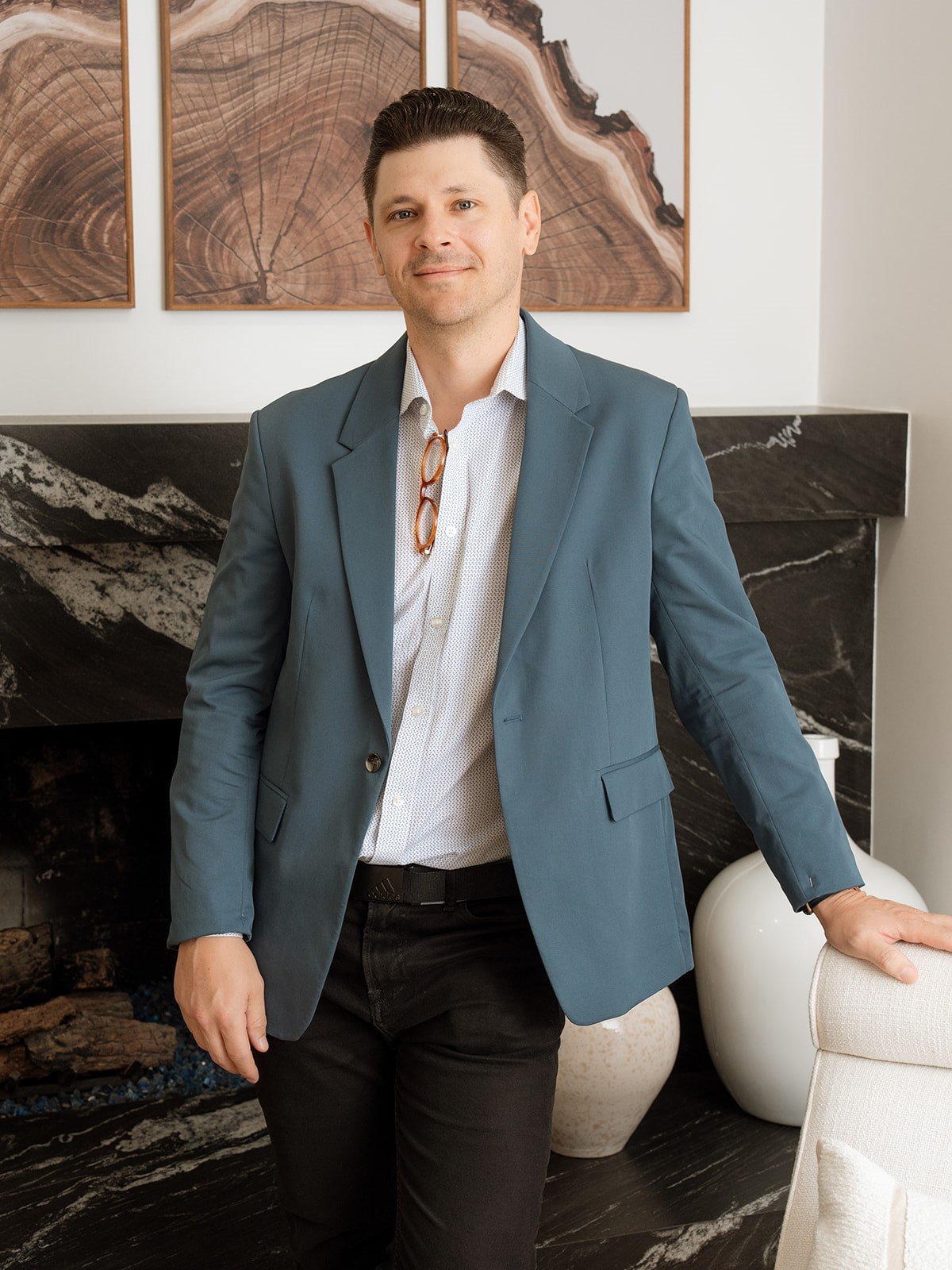 Paul Dement in a blue blazer beside a light upholstered chair, with a stone fireplace behind him.