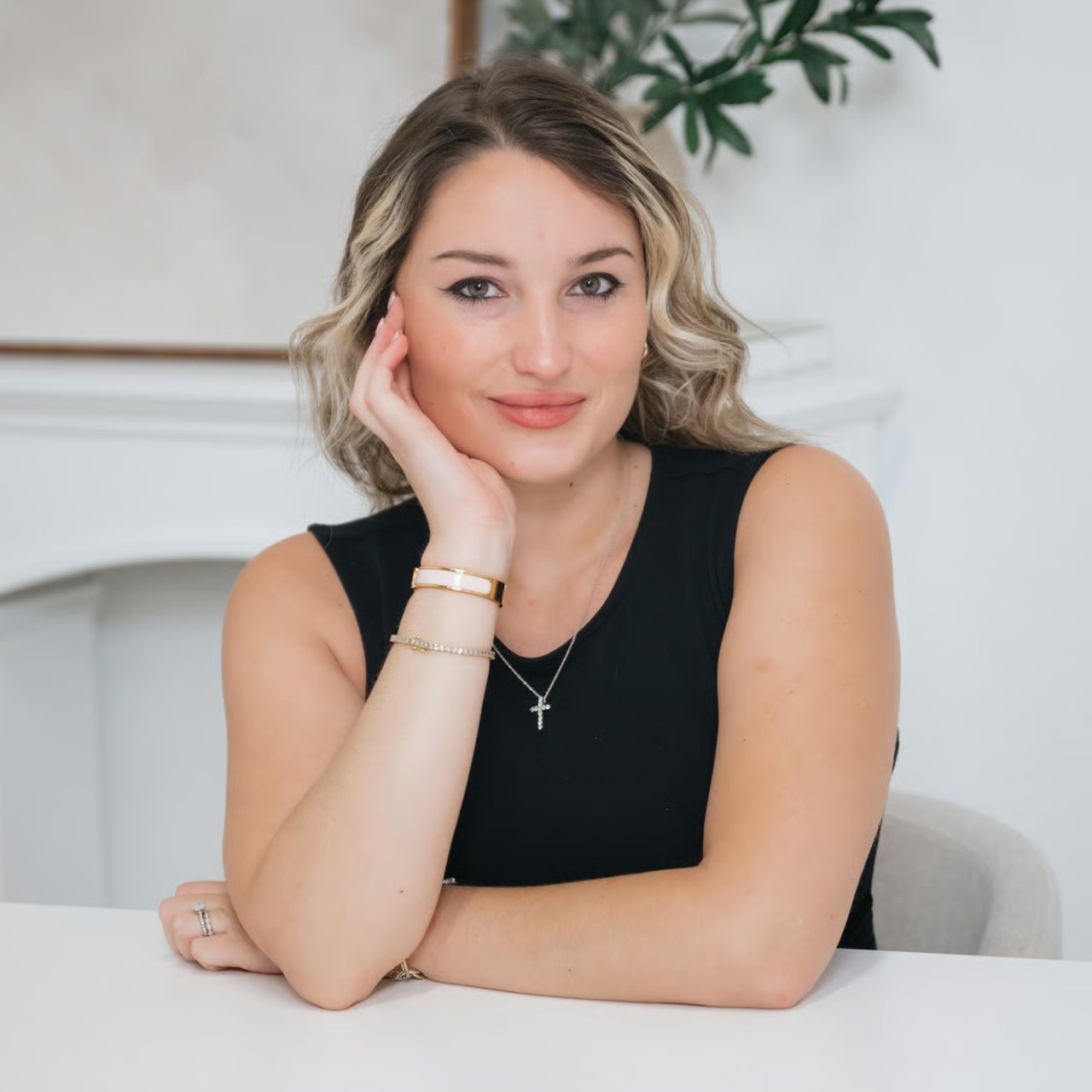 Woman in a black sleeveless top resting her cheek on her hand at a white table.