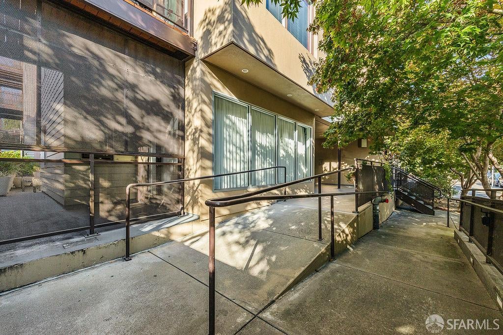 Condominium entrance with a concrete access ramp, metal railings, and leafy trees.