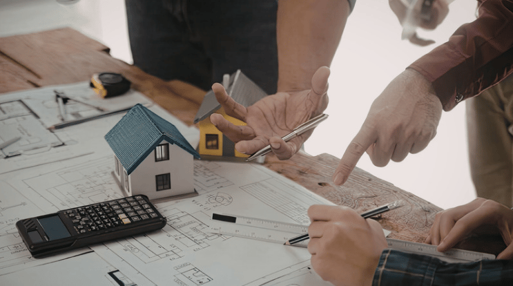 People discuss house plans around a model home and calculator.