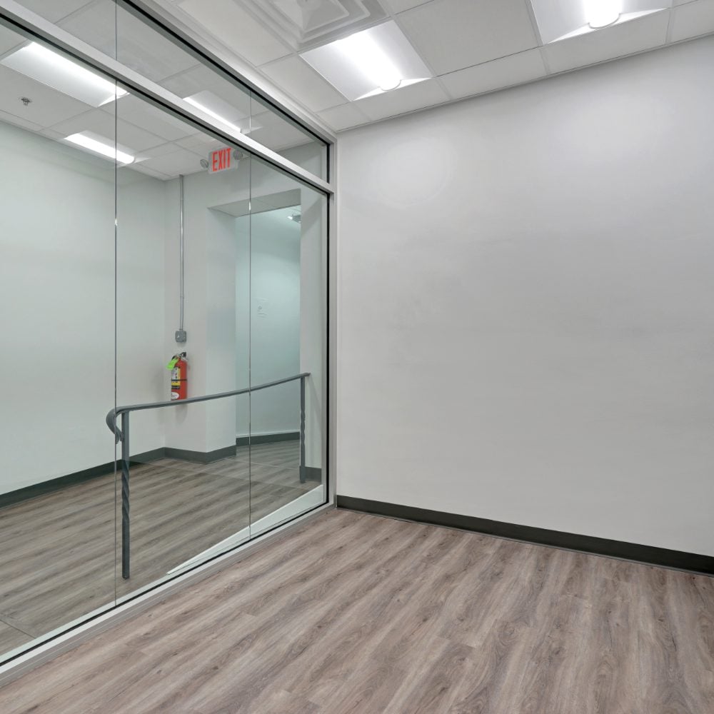 Empty office with a glass partition, gray wood-look flooring, and suspended ceiling.