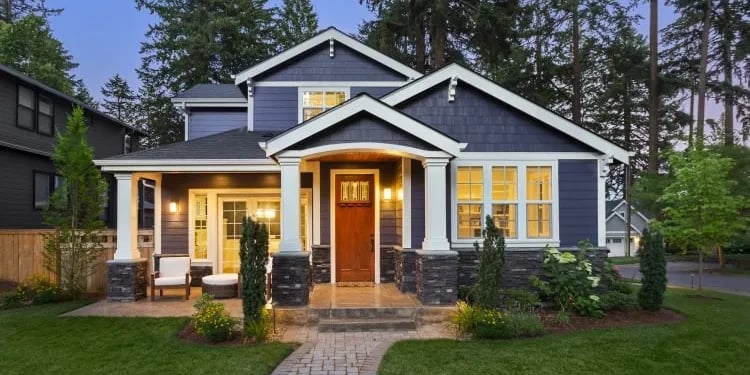 Blue Craftsman home with white-trimmed gables, stone porch columns, and glowing windows at dusk.