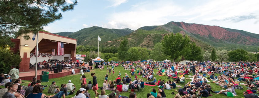 Crowd gathers on the lawn for a concert at a covered stage, with mountains in the background.