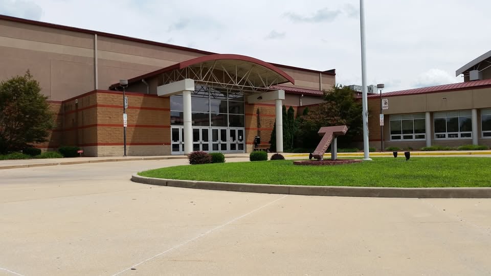 Institutional building with a curved glass entrance canopy and a T-shaped lawn sculpture.