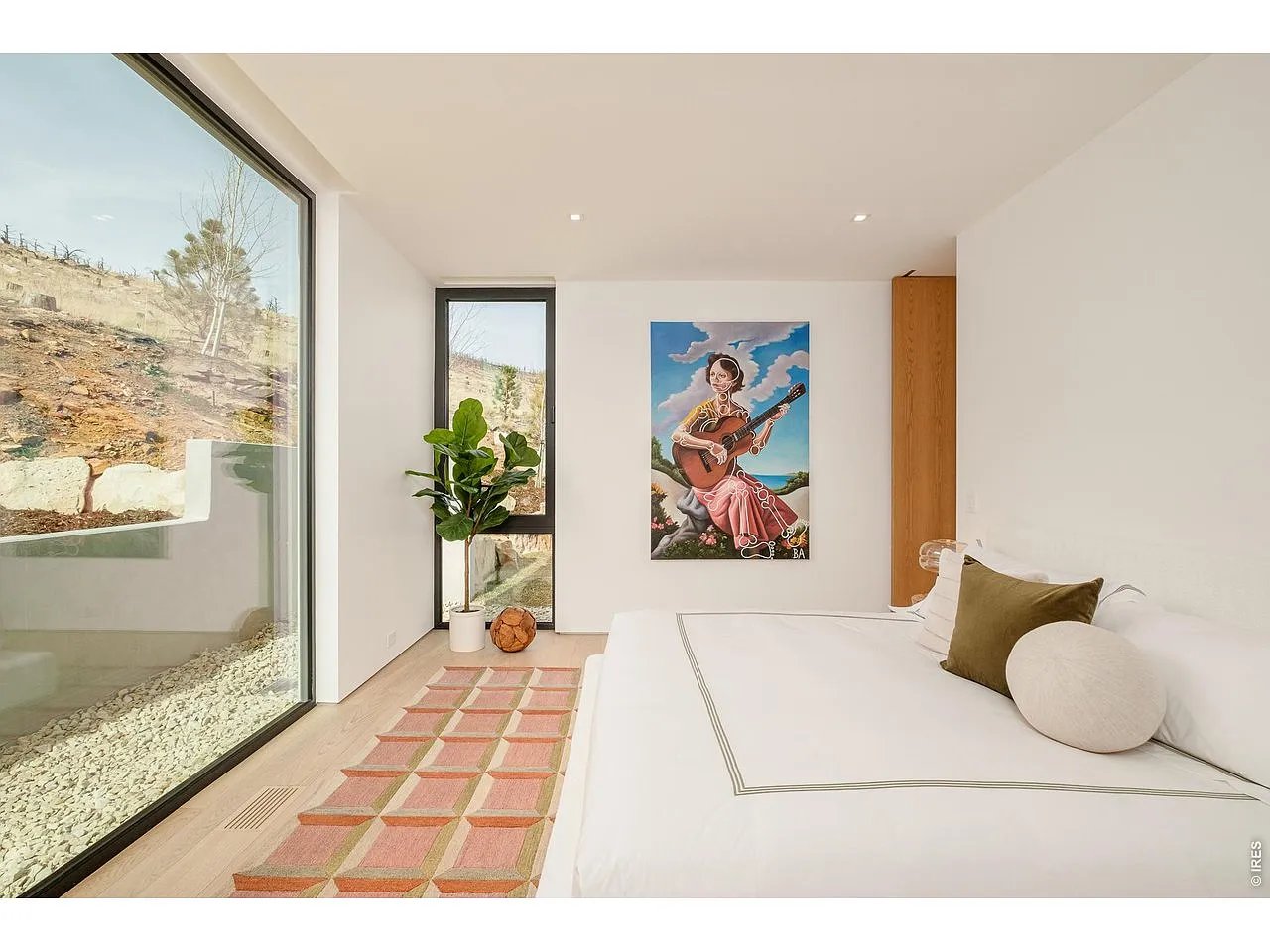 Modern bedroom with a white bed, geometric rug, and large windows overlooking a rocky hillside.
