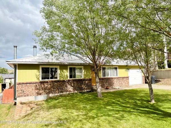 Yellow single-story home with a brick facade, attached garage, and mature trees shading the lawn.