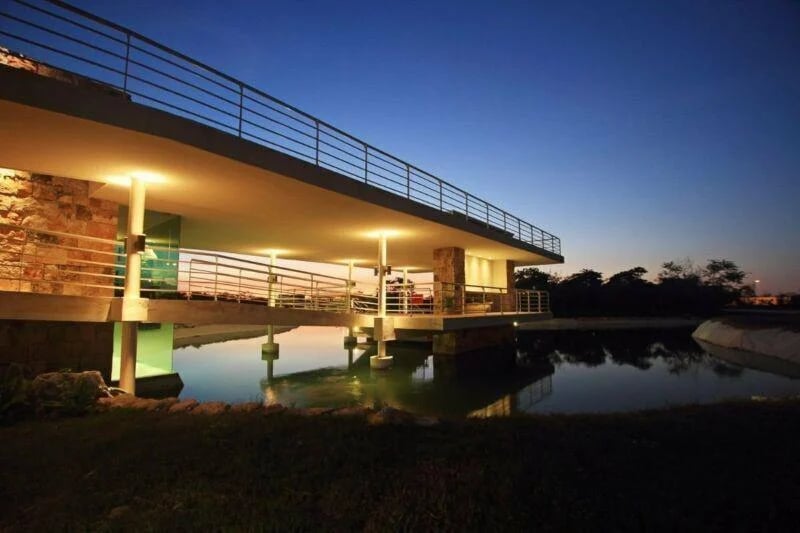 Modern waterfront residence elevated over a pond, with broad terraces and warm lighting at dusk.