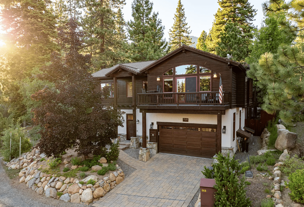 Mountain home with a broad wooden balcony, double garage, and pine trees in warm evening light.