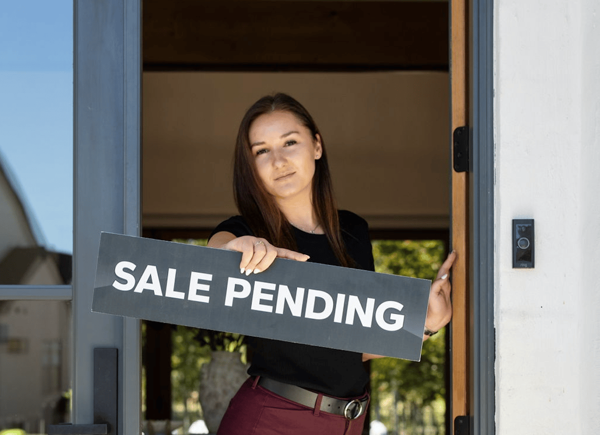 Woman holding a “SALE PENDING” sign in a front doorway.
